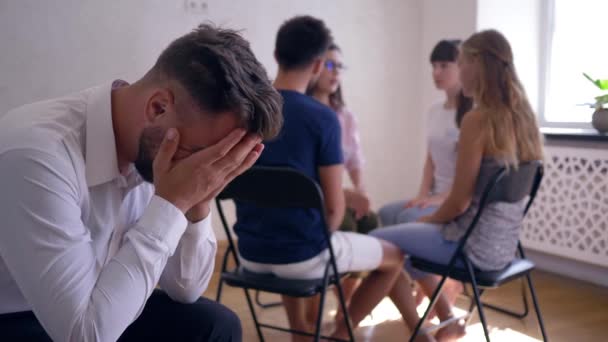 Sad man crying and covers face with hands on group therapy session on background of people sitting on chairs in circle — Stock Video