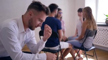 therapy session, sad man thinks about problems on background of people sitting on chairs in circle