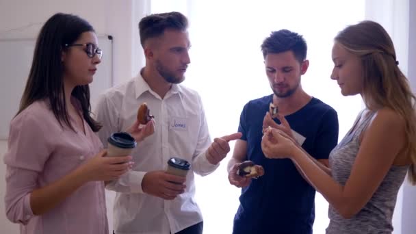 Jovens com crachás seguram pães e café durante o intervalo, trabalhadores de escritório se comunicam e olham na tela do telefone no escritório — Vídeo de Stock