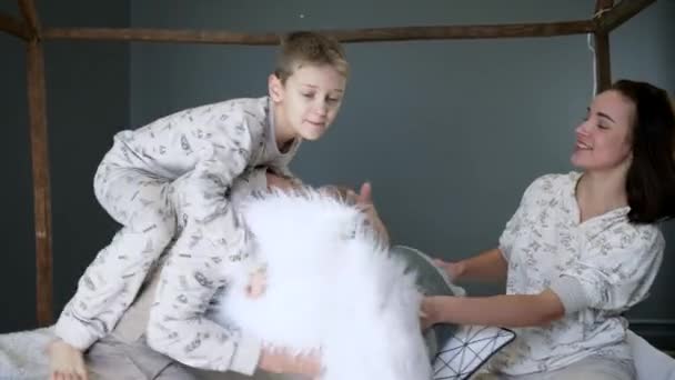 Счастливая семья в одинаковых пижамах бьет друг друга подушками на кровати крупным планом в студии на фотосессии — стоковое видео