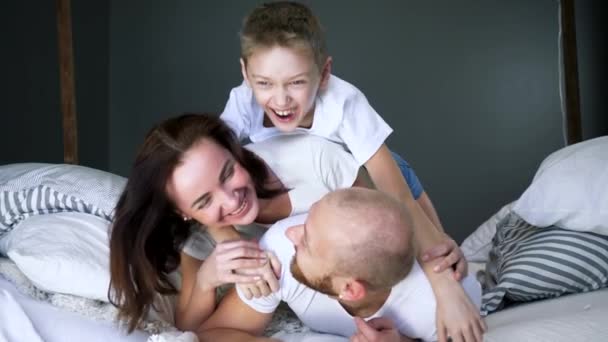 Papá, mamá e hijo en camisetas blancas idénticas se divierten en la cama de cerca en interiores, momentos felices — Vídeos de Stock