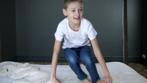Дитинство, хлопчик стрибає на ліжку в повільному русі і дивиться в камеру в спальні — стокове відео