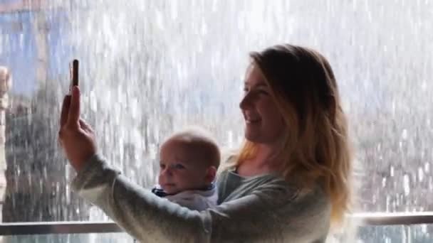 Вихідні, молода мати з дитиною в руках робить селфі фото, використовуючи мобільний телефон на фоні водної стіни — стокове відео