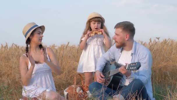 家族の田舎でのレクリエーション若いお父さんがギターを弾くとき、美しい女性が藁帽子と白いドレスで踊り手を拍手し指を叩く一方で、女の子の子供は黄金で甘いパンを食べる — ストック動画