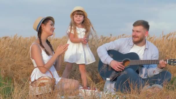 Family fun in nature, young cheerful daddy plays guitar when his beautiful wife and little daugther clap hands happily dancing during picnic in shining by sunbeams wheat harvest field — Stock Video