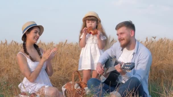 Family outdoor holidays, young daddy plays guitar while his wife claps hands and their little daughter in straw hat and white dress eats sweet baked bun at open air picnic in sunny grain harvest wheat — Stock Video