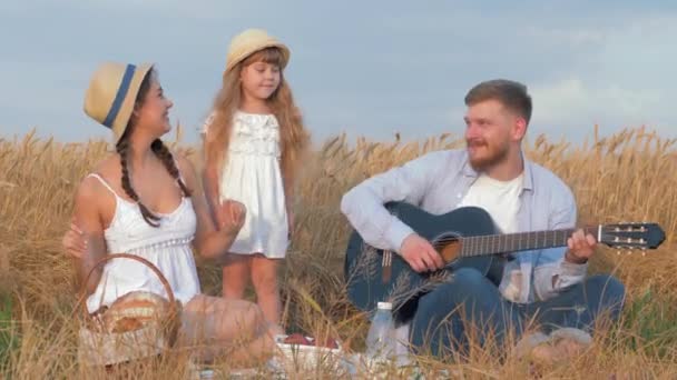 Family idyllic picnic, merry young couple with cute little daughter play musical string instrument and have fun in harvest wheat field shining by autumn sun — Stock Video