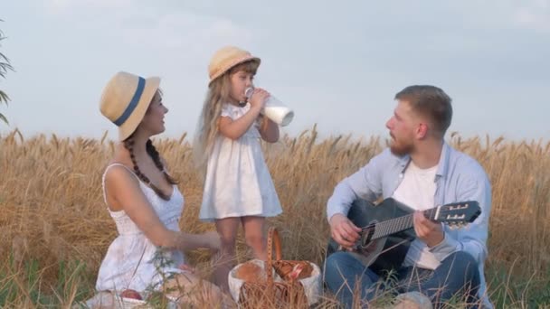 Család vidéken piknik, kislány iszik tejet palack alatt kirándulások a fiatal anya és boldog apa gitározik napsütötte aratott búzamezőn a betakarítás idején — Stock videók
