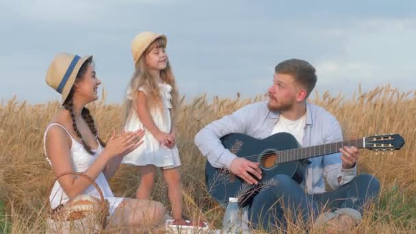 Família ao ar livre diversão, jovem toca guitarra, enquanto sua mulher e pequena filha bonito em chapéus de palha e vestidos brancos palmas mãos regozijando-se no piquenique ao ar livre no campo de cevada grão no tempo de rendimento — Vídeo de Stock