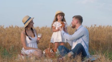 Mutlu aile portresi, genç baba kızının ellerinden tatlı çörekleri ısırır ve güler açık havada güneşli altın rengi çayır pikniğinde oturan kadına güler sonbahar mevsiminde.