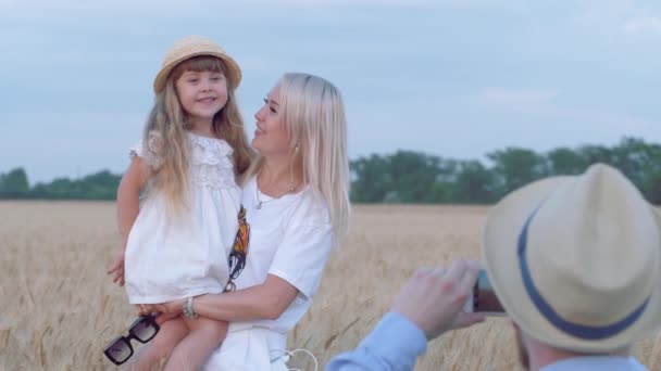 Lycklig familj porträtt, far gör foto på mobiltelefon av kramar och kysser ung mor och liten dotter i höst skörd säsong spannmål havre fält — Stockvideo