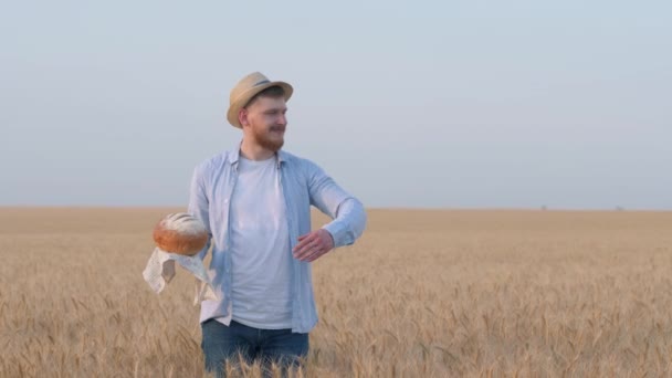 Перспективный молодой агроном, счастливый парень держит хлеб в руке, нюхает его и показывает рукой обширные зерновые поля пшеницы в урожайности осенний сезон против неба — стоковое видео