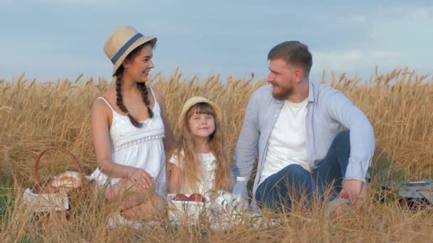 Портрет молодой семьи на открытом воздухе, счастливая пара сидит со своей маленькой дочерью, улыбаясь на пикнике в осенний урожай зерна пшеницы сезонного поля — стоковое видео