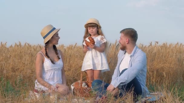 Vidéki családi portré, kis boldog gyerek lány eszik finom édes zsemlét, és kezeli a szüleit a faluban piknik aratott búza termés szezonális mező — Stock videók