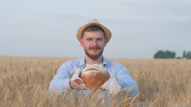 Joven cultivador de grano feliz, hombre guapo en sombrero de paja le da a la cámara sabroso pan horneado y sonrisas de pie en el campo de picos de avena durante la temporada de rendimiento — Vídeos de Stock