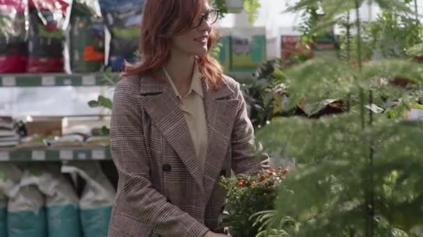 Attractive woman in glasses for sight florist holds in her hands blooming flower in pot background of green plants in department store for gardeners — Stock Video