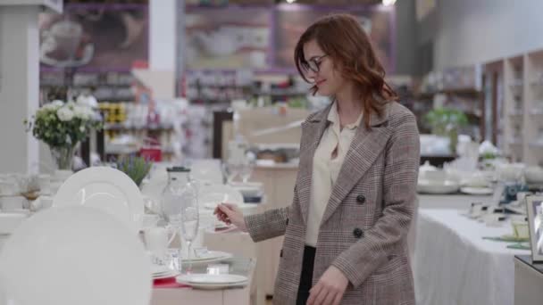 Приваблива домогосподарка в окулярах для бачення вибирає сучасні страви для набору посуду в магазині обладнання — стокове відео