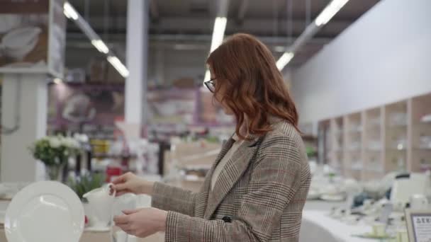 Jovem comprador feliz fêmea escolhe conjunto de chá para seu apartamento na loja com pratos, bela mulher em óculos para os olhos examinados pratos brancos no supermercado — Vídeo de Stock