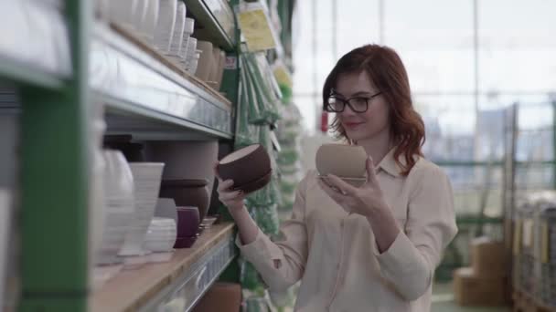 Décor et concept de design, femme souriante dans des lunettes pour la vision choisit des pots de couleur modernes pour les plantes à la maison pour le design intérieur de la maison ou du bureau dans un grand magasin de plantes vertes — Video