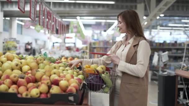 Minorista, joven compradora con cesta de comida en las manos recoge fruta fresca de manzana en el supermercado, sonríe mira a la cámara y muestra el cartel de la clase con su mano — Vídeos de Stock
