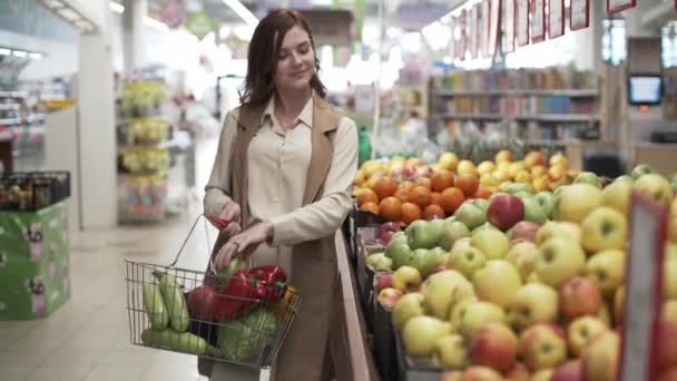 Πορτρέτο μιας όμορφης γυναίκας αγοραστή με καλάθι για προϊόντα στα χέρια της που επιλέγει φρέσκα φρούτα και λαχανικά σε μια αγορά σούπερ μάρκετ — Αρχείο Βίντεο
