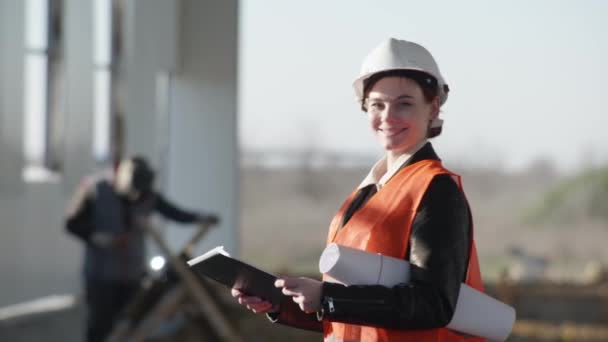Női mérnök védősisakban rajzokkal a kezében a háttérben az ember hegesztő gép és szikrák építése során a hangár, építés — Stock videók