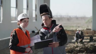 Koruyucu giysili erkek ve kadın mühendisler, güvenlik kaskları, inşaat alanındaki hangar tasarımı ve ofis çizimlerini kontrol ediyorlar.