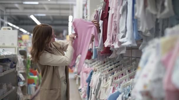 Gelukkige jonge vrouw kiezen kleding voor kinderen, mooie vrouw in de winkel proberen kwaliteit van kleding, baby goederen winkel — Stockvideo