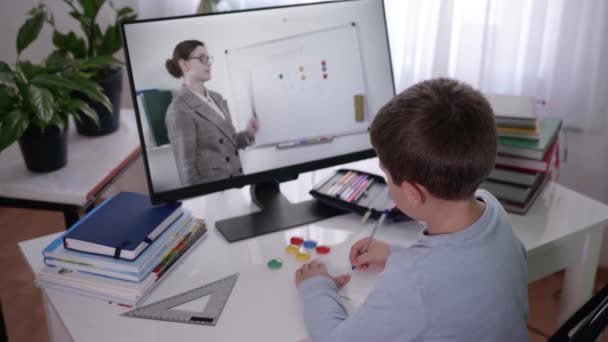 Educación en línea, estudiante con libros de texto en la mesa en el estudio en casa hace la tarea con la ayuda de las tecnologías modernas para los niños viendo lección remota con el maestro — Vídeo de stock