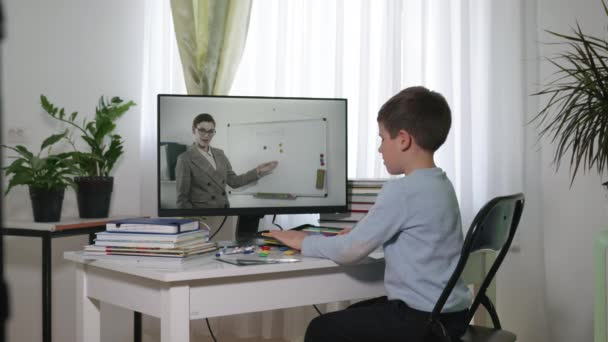 Scuola on-line, ragazzo impara utilizzando lezioni video con insegnante, guarda schermo del monitor e risponde compiti mentre seduto a casa a tavola con i libri durante l'isolamento e la quarantena — Video Stock