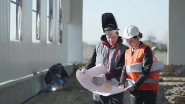 Koruyucu miğferli kadın mimar, arkaplanda profesyonel bir işçiyle birlikte çizimlere göre hangarın inşaatını tartışıyor.