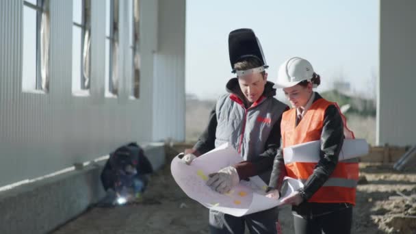 Female architect in protective helmet discusses the construction work of hangar according to drawings with a professional employee on background of welding — Stock Video