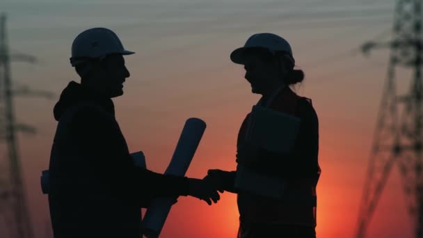 A mulher de empregado de engenheiros e o homem em capacetes protetores e coletes discutem a estrutura ou a reconstrução de novas linhas elétricas modernas de cidades poder de alta tensão em um campo contra um pôr-do-sol, silhueta — Vídeo de Stock