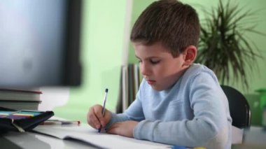 Küçük akıllı ilkokul öğrencisi evde bilgisayar başında oturup öğretmeniyle birlikte modern ekranda ders videosu izleyerek ders öğreniyor.