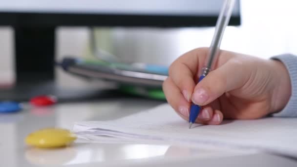 Niño escribe números con pluma y ejemplos de matemáticas en cuaderno sentado en el fondo de la mesa de útiles escolares, manos de cerca — Vídeo de stock