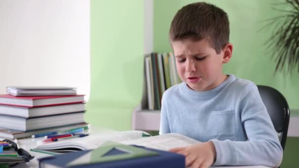 Educação em casa, criança cansada chateada faz lição de casa, lê livro e sustenta sua testa com a mão enquanto se senta à mesa com material escolar, aprendizagem difícil — Vídeo de Stock