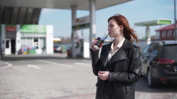 Jó szolgáltatás, mosolygós lány csésze kávé magával áll a háttérben a benzinkút, míg az autó tele van benzin, benzin árak koncepció — Stock videók
