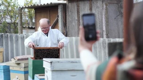 Apicultor velho moderno com a ajuda de sua esposa filma um vídeo no smartphone para vlog e mostra assinantes favos de mel no fundo de colmeias e abelhas — Vídeo de Stock