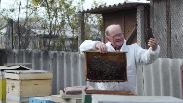 Apiculture, influenceur masculin âgé en costume de protection avec des lunettes de vue fait blog vidéo pour les adeptes avec téléphone portable dans les mains montre nid d'abeilles sur fond de miel debout dans un — Video