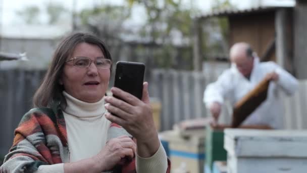 Narablog wanita tua dengan smartphone di tangannya berbicara melalui link video tentang peternakan lebah yang menunjukkan seorang pria dalam setelan pelindung dengan sarang lebah di tangannya latar belakang sarang lebah madu dan lebah madu — Stok Video