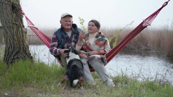 Mooie romantische middelbare leeftijd oudere grootouders ontspannen plezier samen zitten in hangmat in de buurt van rivier genieten zorg tederheid — Stockvideo