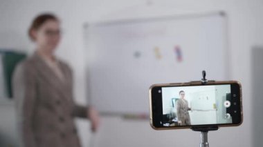 Çevrimiçi eğitim, vizyon gözlüklü modern kadın pedagog karantina sırasında beyaz bir tahtaya karşı akıllı telefon kullanan ilkokul öğrencilerine ev ödevi anlatıyor