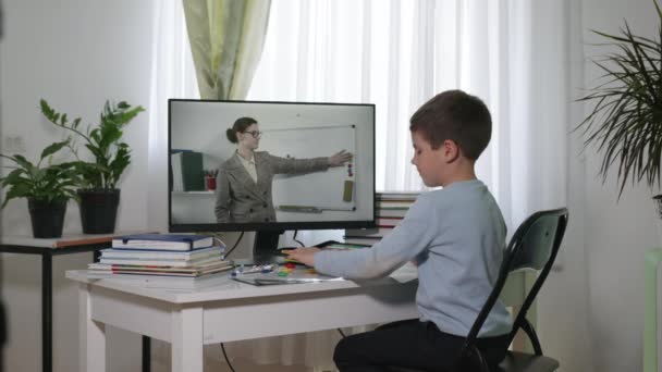 Niño estudiante de primaria viendo una videolección en línea en la pantalla de un monitor mientras está sentado en casa en una mesa con útiles escolares, pandemia — Vídeos de Stock