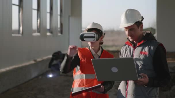 Modern teknik, kvinnlig ingenjör i skyddshjälm och viar glasögon tittar på modell av ny hangar tillsammans med manlig kollega i hjälm och laptop i händerna — Stockvideo
