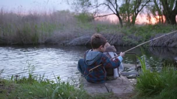 Маленькие рыбаки сидят на причале и ловят рыбу с деревянной удочкой в ручье посреди заката во время веселого отдыха за городом среди тростника. — стоковое видео