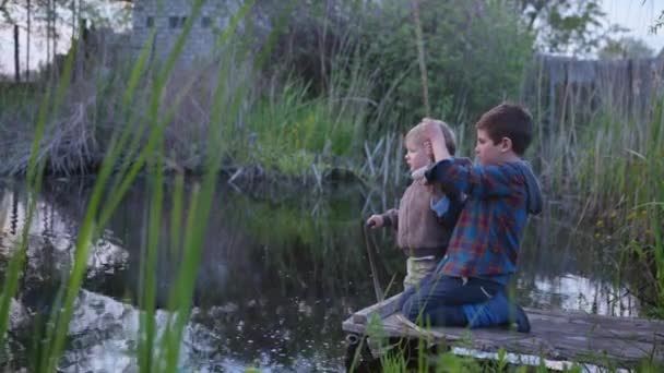 Gelukkige jeugd, actieve kinderen, jongens vangen vis op een houten hengel zittend op een pier bij de rivier in het midden van riet op een warme dag — Stockvideo