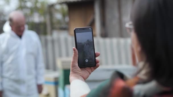 身穿防护服的有影响力的男性养蜂人为博客拍摄视频，并在他与妻子的手机上告诉他的追随者蜂群和蜂群中的蜂群 — 图库视频影像