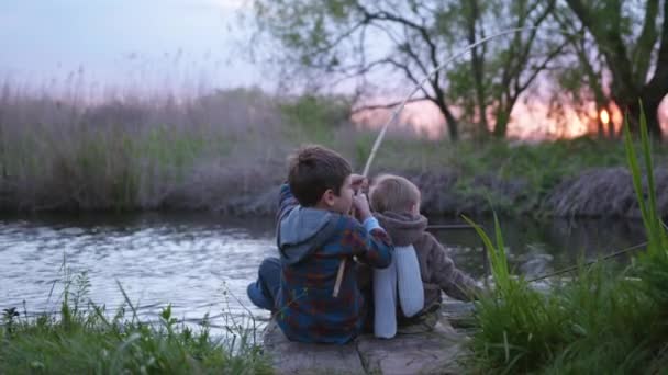 Familie kleine visser, kinderen zitten op pier en vis met een hengel in het meer bij zonsondergang op buiten tussen riet, achteraanzicht — Stockvideo
