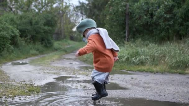 Παιχνίδι υπαίθριο, μικρό παιδί αρσενικό σε καπέλο και λαστιχένιες μπότες παίζει σε λακκούβα σε εξωτερικούς χώρους — Αρχείο Βίντεο