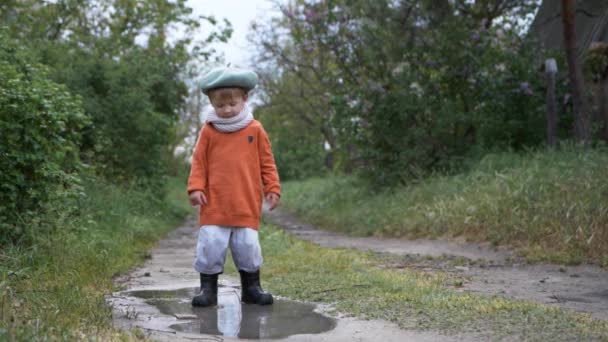 Vacaciones de primavera, lindo niño masculino en sombrero y botas de goma se divierten saltando en charco sucio — Vídeos de Stock
